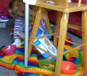 Child hiding under a chair eating crackers from the box.